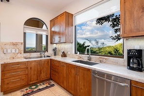 A kitchen with wooden cabinets, a stainless steel dishwasher, a coffee machine, and a large window with a view of greenery.