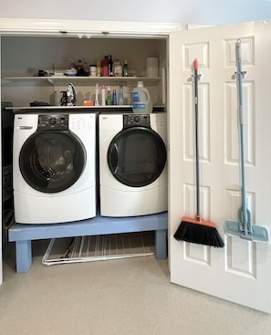 A laundry area with two front-loading washing machines, a shelf with cleaning supplies, and a broom leaning against the wall.
