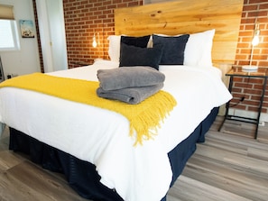 A hotel room with a brick wall, a wooden headboard, a bed with white bedding, a yellow throw, and a small table with a lamp. - Your Boulder Home Base, Walk to Pearl St, 2BR/2BA + Parking! (Boulder)