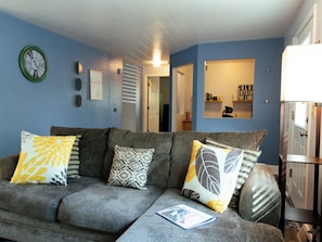 A living room with a grey sofa, patterned cushions, a wall clock, and a bookshelf.