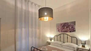 A neatly arranged bedroom with a bed, bedside lamps, a hanging light fixture, and a wall-mounted artwork. - Gracchi 278 - The Italian Stay Collection (Rome)