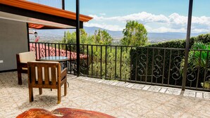 A balcony with a patterned floor, a wooden chair, a table, and a view of the city and mountains. - Room just 20 minutes from the airport. (Alajuela)