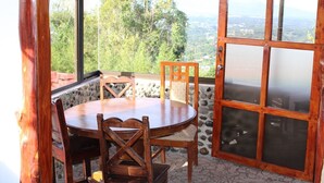 A wooden dining table with chairs in a room with a stone wall and large windows offering a view of trees and hills. - Room just 20 minutes from the airport. (Alajuela)