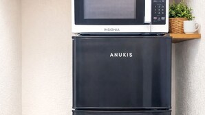 Un refrigerador Anukis negro con un microondas encima, ubicado en un rincón con piso de madera y pared blanca con textura.