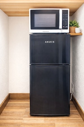 A black Anukis refrigerator with a microwave on top, placed in a corner with a wooden floor and a white textured wall. - Barrel Cactus Container Home (Azle)