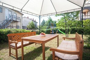 Un patio exterior con una mesa y bancos de madera, una zona de juegos con un columpio y una zona cubierta con un toldo blanco.