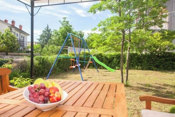 A wooden table with a bowl of fruit and a swing set in a garden.