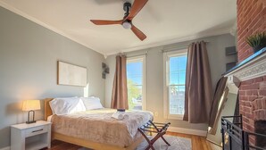 Un dormitorio con cama, mesita de noche, ventilador de techo, ventana con cortinas y chimenea de ladrillo.