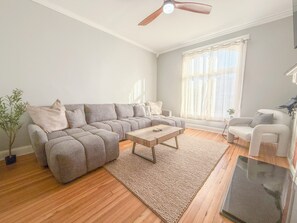 Uma sala de estar com sofá seccional, uma mesa de centro, ventilador de teto e uma grande janela com cortinas leves.