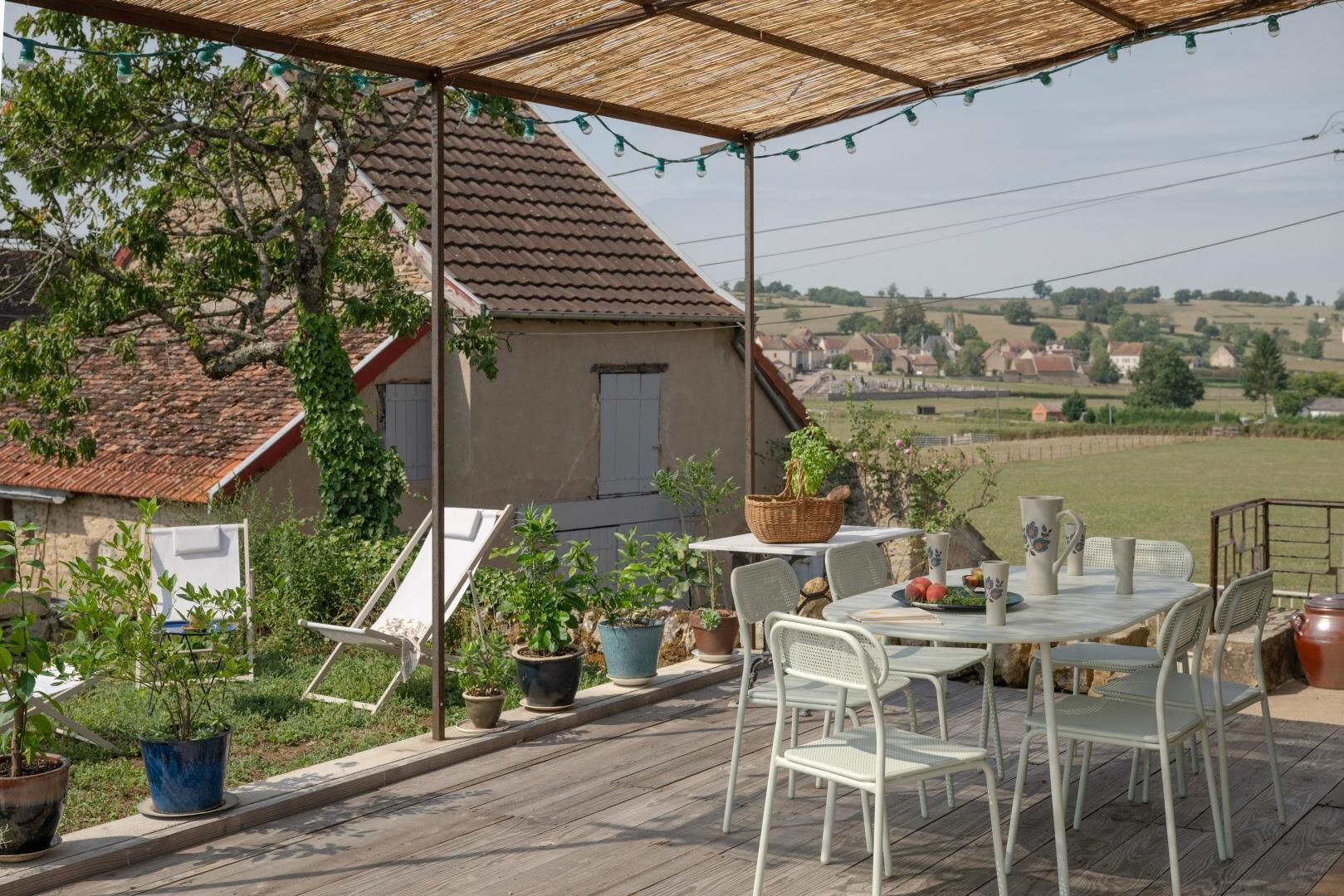 An outdoor patio with a table and chairs, surrounded by potted plants and a view of a rural landscape.