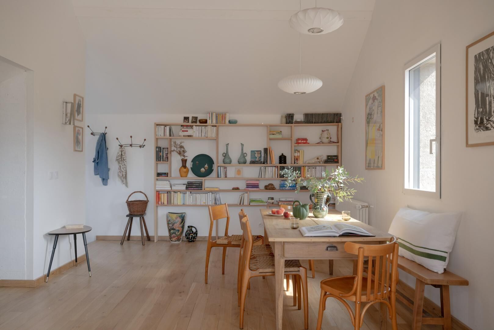 A spacious room with a wooden floor, a dining table with chairs, a bookshelf filled with books and decorative items, and a large window providing natural light.