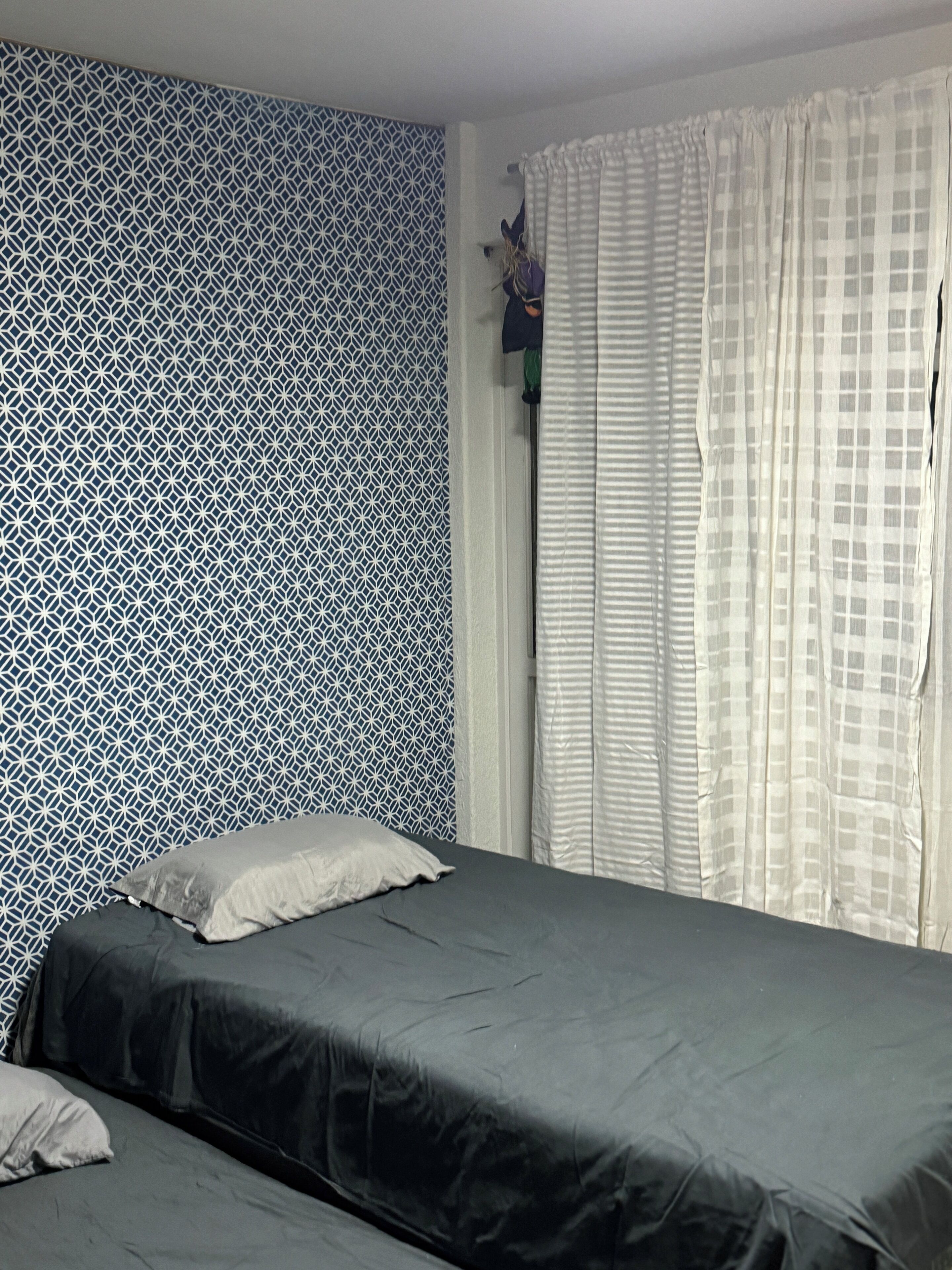 Una habitación con pared estampada, cama con cubrecolcha oscuro y ventana con cortinas blancas.