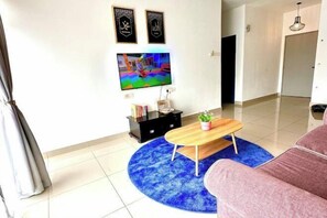 Ein Wohnzimmer mit Flachbildfernseher, Couchtisch aus Holz, blauem Teppich, rosa Sofa und zwei gerahmten Bildern an der Wand.