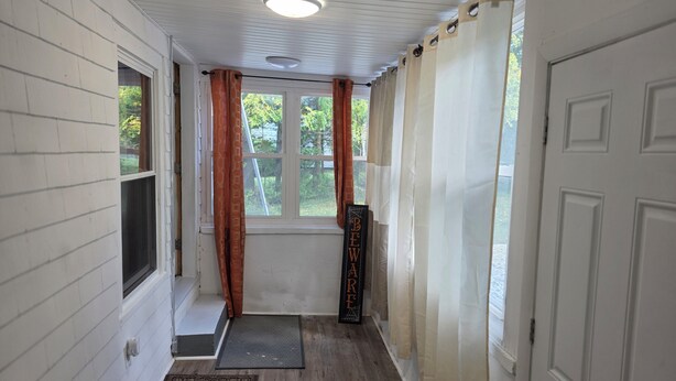 A hallway with white walls, a wooden floor, a window with orange curtains, and a door with a nameplate.