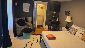 A bedroom with a bed, a desk, a chair, a lamp, and a door leading to another room.