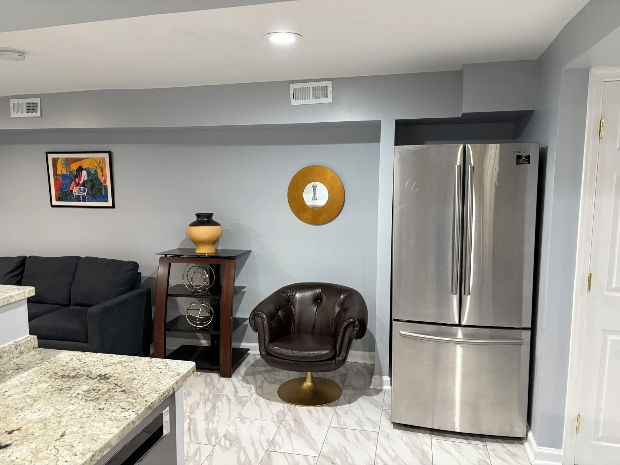 A modern kitchen with a marble countertop, a black leather chair, a stainless steel refrigerator, and a wall-mounted clock.