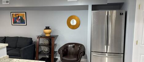 A modern kitchen with a marble countertop, a black leather chair, a stainless steel refrigerator, and a wall-mounted clock.