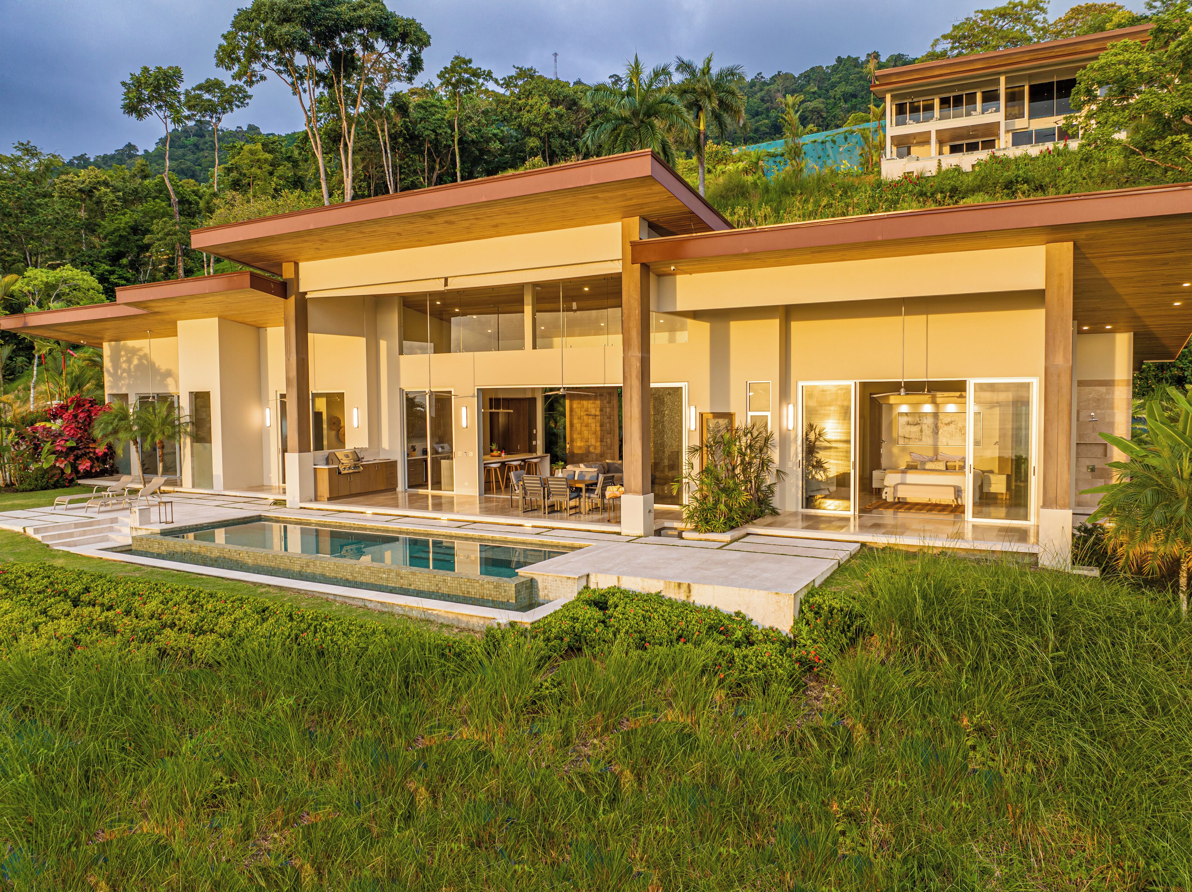 Une maison moderne à deux étages, dotée d’une piscine, de grandes fenêtres et d’un jardin luxuriant.