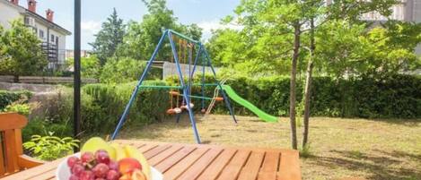 A wooden table with a bowl of fruit and a swing set in a garden.
