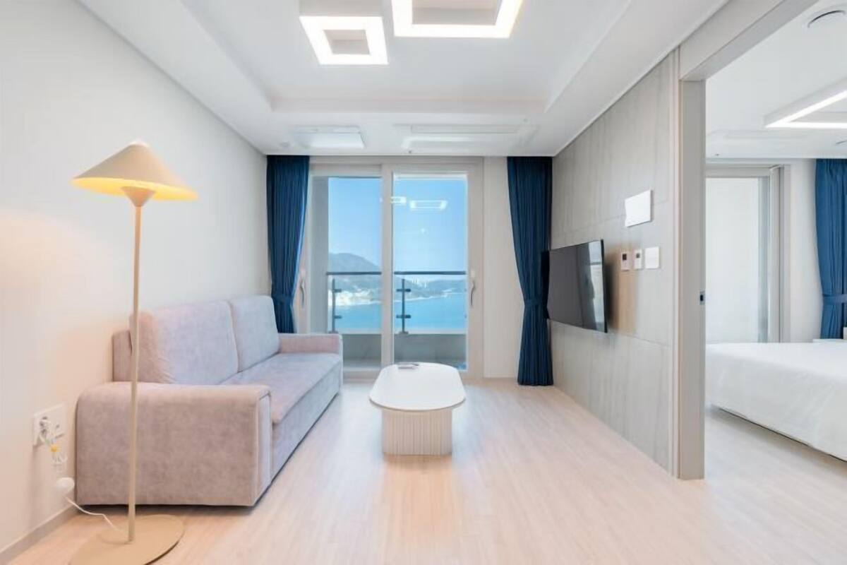 A modern hotel room with a sofa, a coffee table, a bed, a TV, and a balcony with a view.