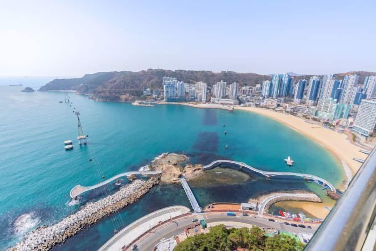A coastal cityscape with a clear view of a beach, a rocky breakwater, and high-rise buildings.