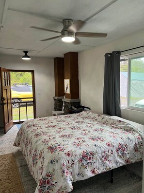 A bedroom with a bed, a ceiling fan, a window with curtains, a door leading outside, and a small table with a chair. - Your Cozy Aloha Room (Royal Hawaiian Estates)