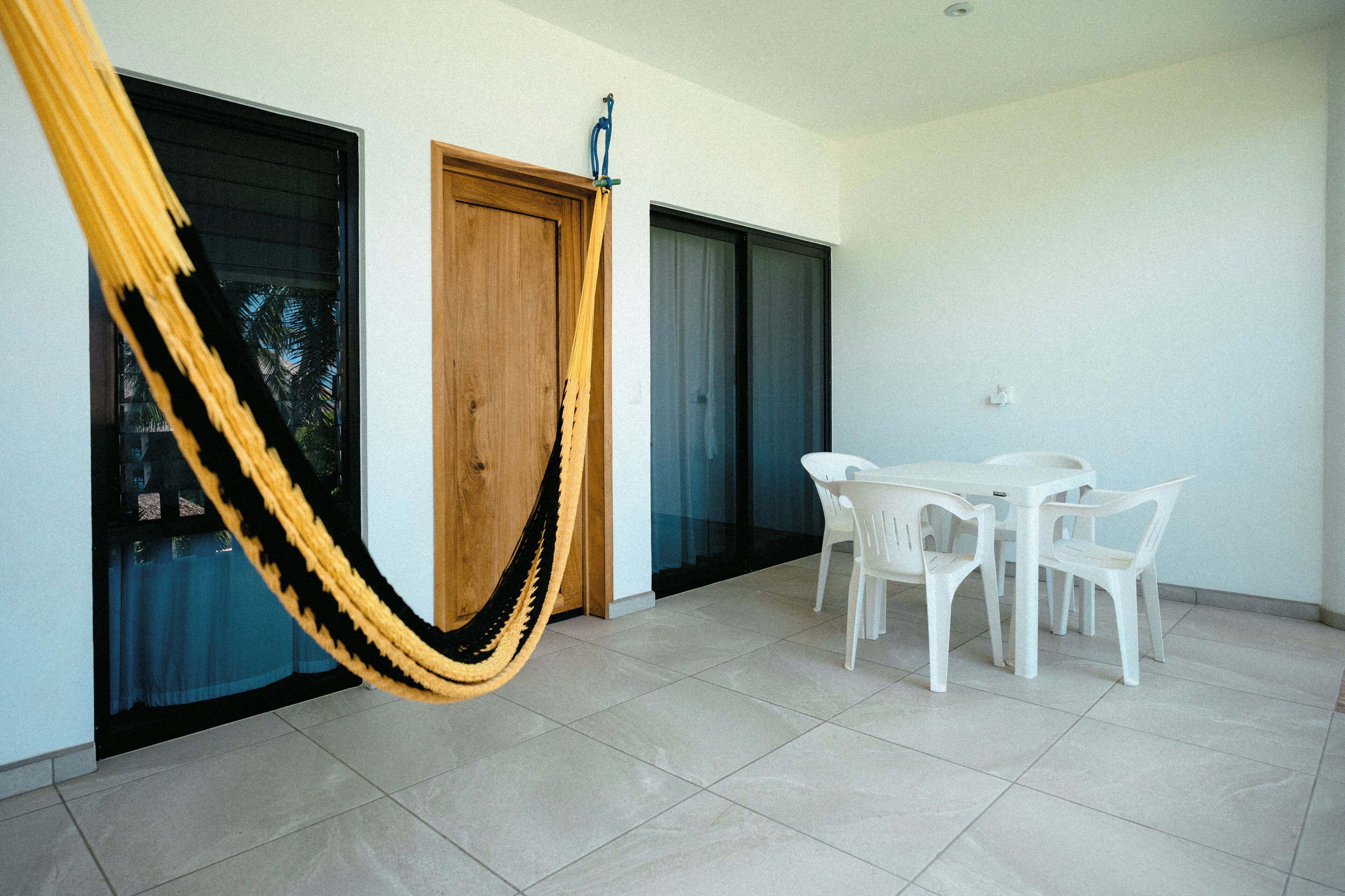 Una habitación con una hamaca, una puerta de madera, una puerta de cristal, una mesa blanca con sillas y un piso de baldosas.
