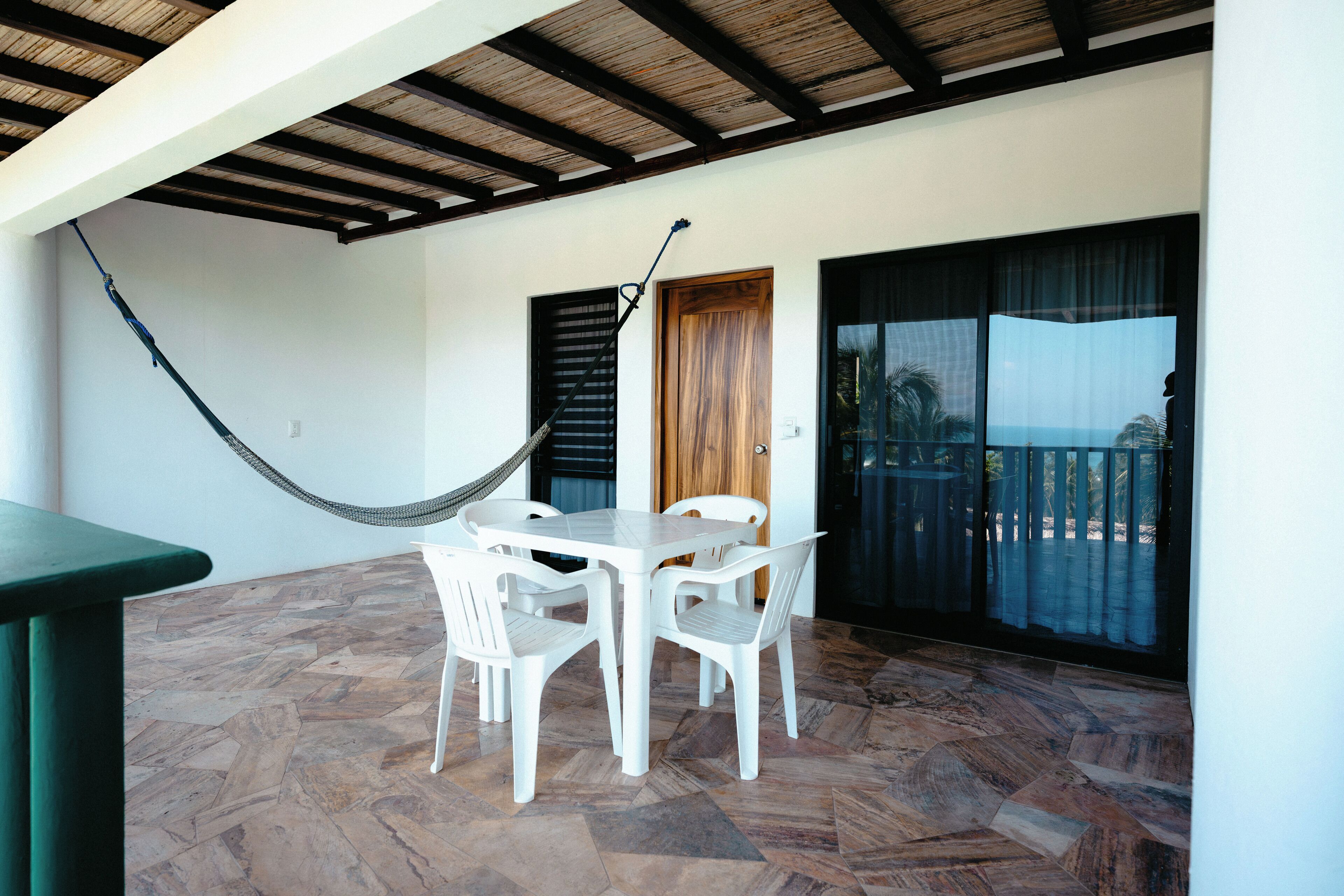 A room with a wooden ceiling, a hammock, white plastic chairs, a table, and a view through large windows.