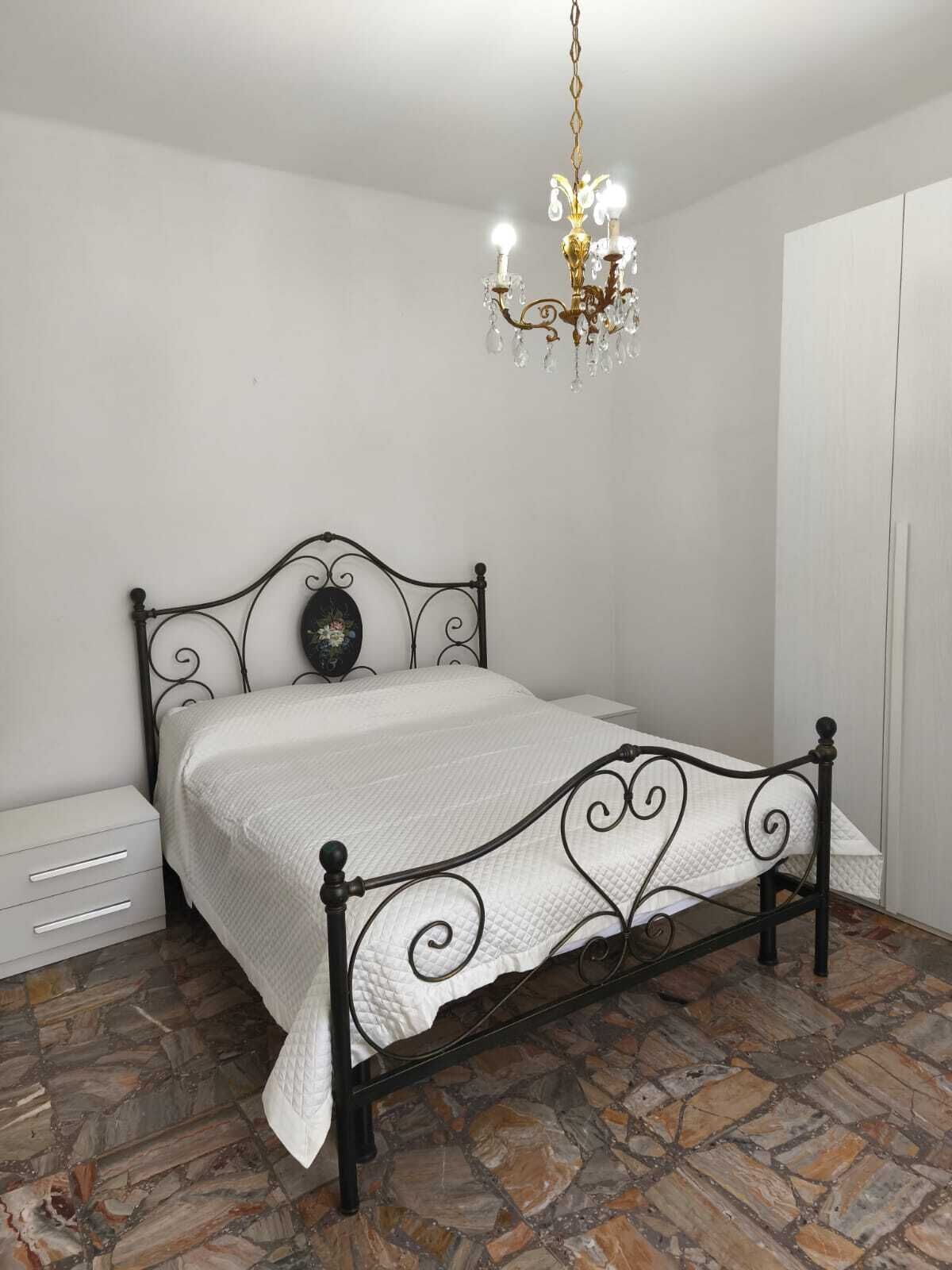 Una camera da letto con un letto a struttura in metallo, biancheria da letto bianca, un lampadario e un pavimento in pietra.