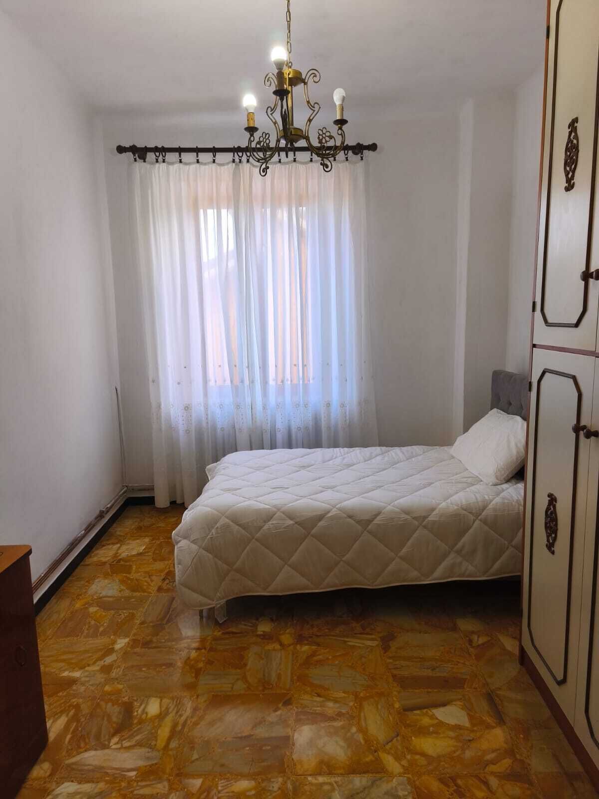 A bedroom with a bed, a chandelier, a wardrobe, and a window with curtains.