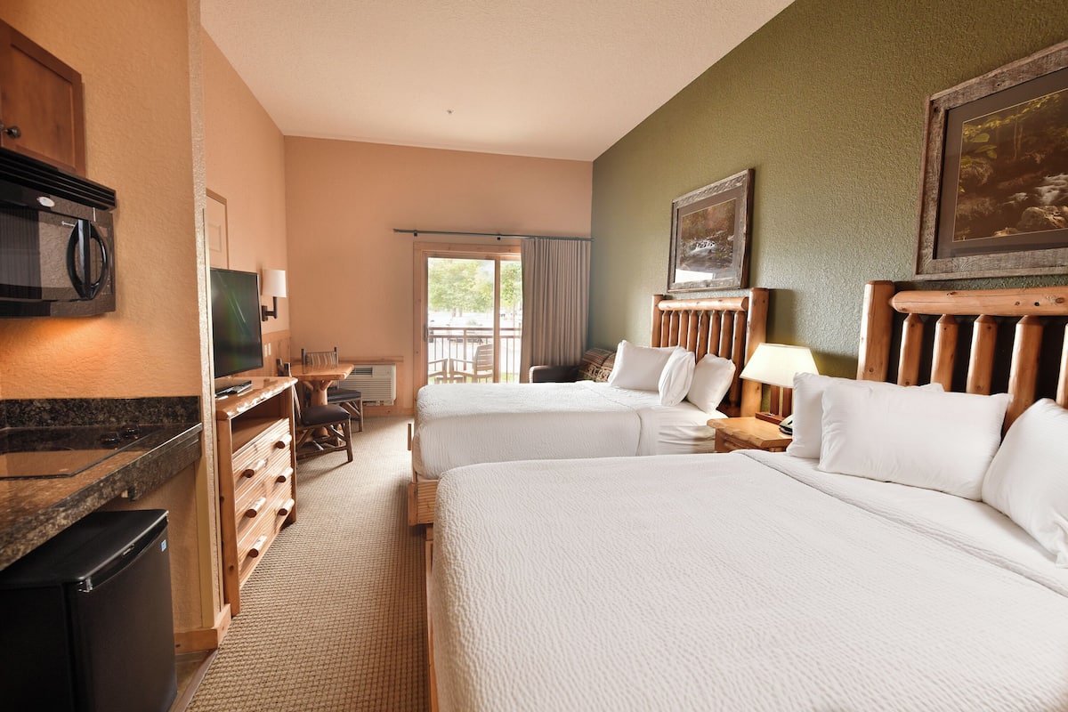 A hotel room with two beds, a kitchenette, a television, and a view of the outdoors.