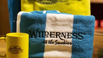 Una persona con una camiseta azul con el logo 'RUMAWAY CANYON', una toalla amarilla, una toalla azul y blanca, y una taza amarilla con el logo 'WILDERNESS at the Smokies'.