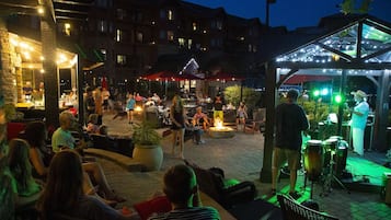 An outdoor evening gathering with people seated and standing around a central fire feature, with a band performing on a stage.