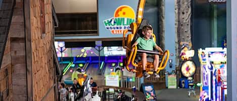 A child on a swing ride at an indoor amusement park with various arcade games and a sign for "HOWLIN' HOUND DOOR".