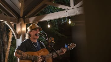 Eine Person spielt eine Akustikgitarre in einem überdachten Außenbereich mit Holzbalken und Lichterketten.