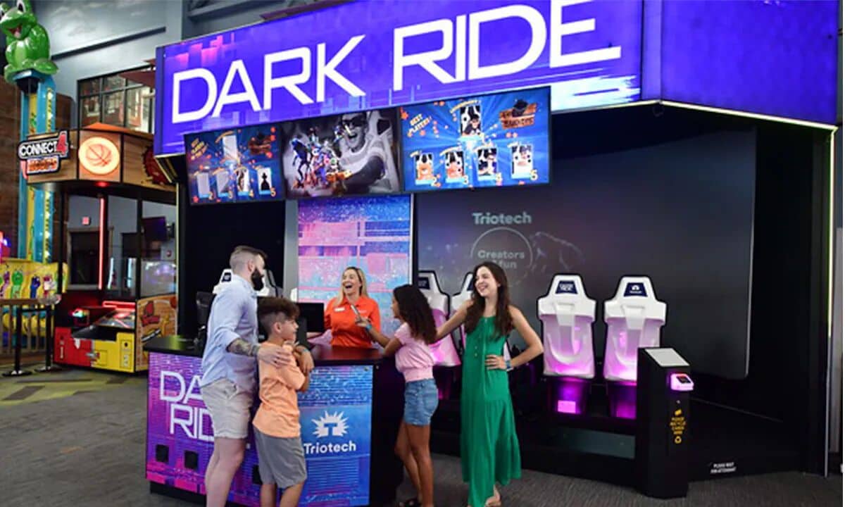 A family interacting with a 'Dark Ride' attraction booth with a 'Triotech' logo, featuring a digital display and a 'Connect4' game machine.