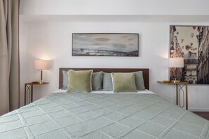 A hotel room with a bed, two bedside lamps, a painting above the bed, and a view of a cityscape through the window.