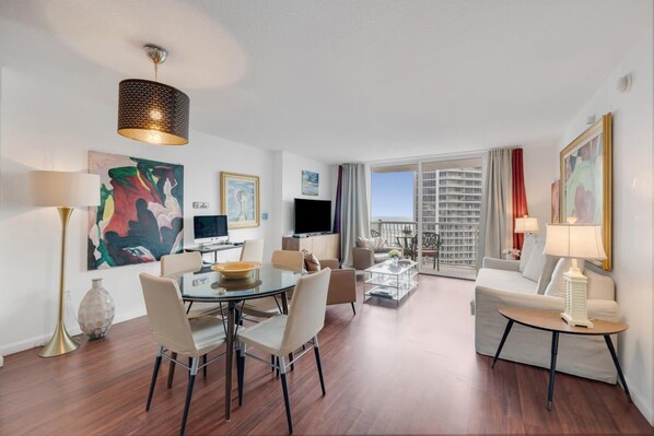 A modern hotel room with a dining area, a living area with a sofa and a TV, and a balcony with a city view.