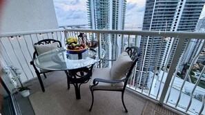 A balcony with a glass table and chairs, a fruit bowl, and a view of high-rise buildings.