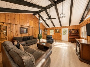 A spacious living room with a high ceiling, wooden walls, a leather sofa, a green armchair, a piano, and a television. - Rustic Lodge • Pet-Friendly Cabin w/ Hot Tub & Fire Pit (Pigeon Forge)