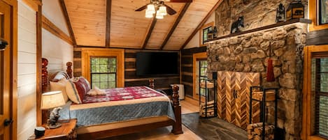 Un dormitorio tipo cabaña de troncos con una cama, una chimenea de piedra, un televisor y un ventilador de techo.
