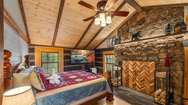 Une chambre confortable dans un chalet en rondins, avec un grand lit, une cheminée en pierre et un téléviseur fixé au mur.