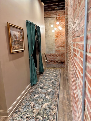 Un pasillo con alfombra estampada, un cuadro enmarcado, cortinas verdes y una pared de ladrillo.