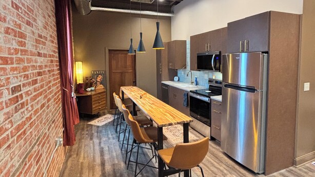 A modern kitchen with a long wooden table, four chairs, stainless steel appliances, and exposed brick walls.