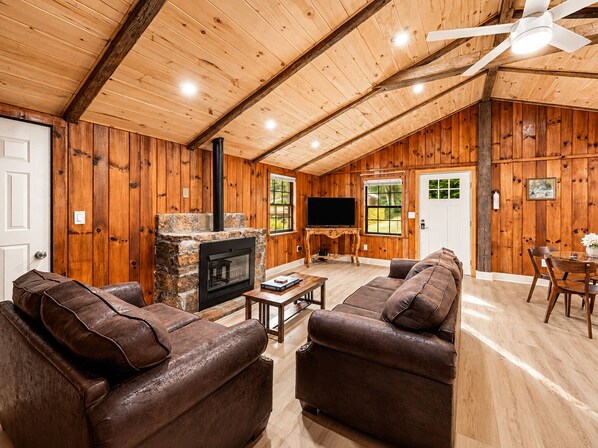 A log cabin interior with a stone fireplace, leather sofas, a television, and a dining area.