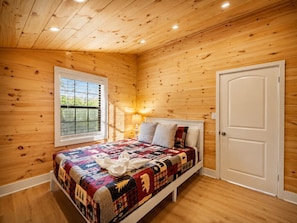 A bedroom with a wooden ceiling, a window with blinds, a bed with a quilt, a bedside lamp, and a door.