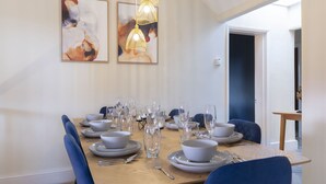 A dining area with a wooden table set for six, surrounded by blue chairs. There are two abstract paintings on the wall above the table.