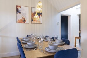 A dining area with a wooden table set for six, surrounded by blue chairs. There are two abstract paintings on the wall above the table.