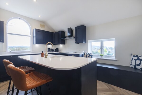 A modern kitchen with dark cabinetry, a large island with bar stools, a built-in oven, and a window providing a view of the city.