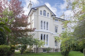 Ein großes, cremefarbenes Gebäude mit zahlreichen Fenstern und einer symmetrischen Fassade, umgeben von üppigem Grün und Bäumen.
